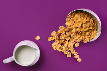 Bowl with corn flakes, jug of milk on purple background, top view