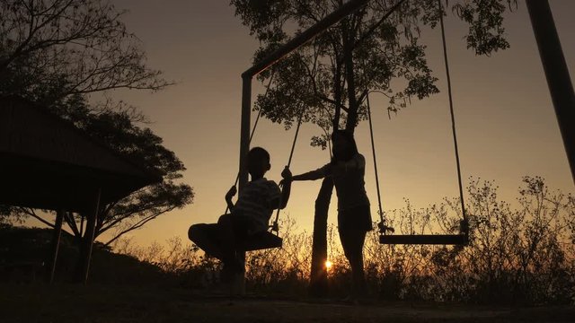 Silhouetted Young Mother With Boy Enjoying Beautiful Sunny Day, Swinging On Swing Set. Red Orange Sunset Background.
