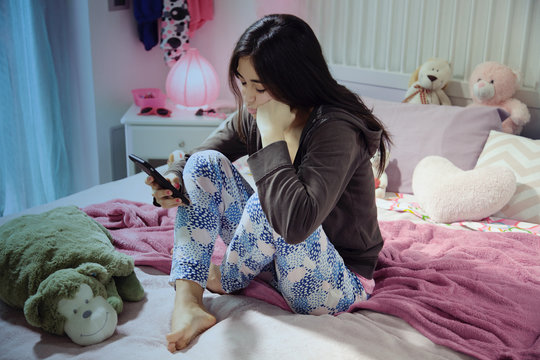Sad hispanic teenager sitting on bed looking message on phone