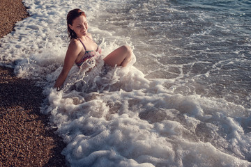 Fototapeta premium Young beautiful smiling woman with red hair in blue and pink swimsuit is sitting on the beach of caribbean sea with at sunset