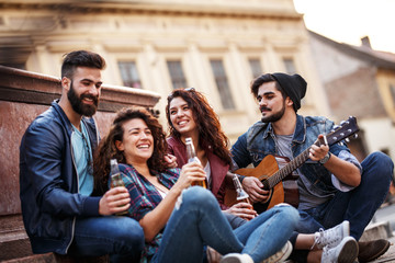 Group of young friends hangout on city square .They playing guitar and sings.