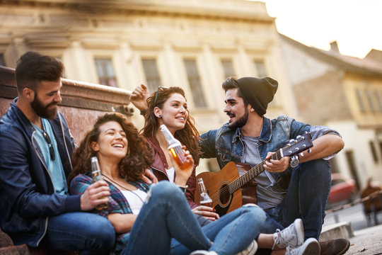 Group of young friends hangout on city square .They playing guitar and sings.