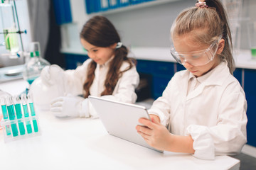 Two little kids in lab coat learning chemistry in school laboratory. Young scientists in protective glasses making experiment in lab or chemical cabinet. Working on a tablet.