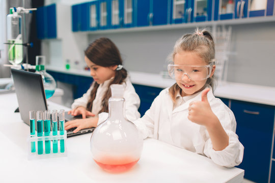 Two little kids in lab coat learning chemistry in school laboratory. Young scientists in protective glasses making experiment in lab or chemical cabinet. thumbs up