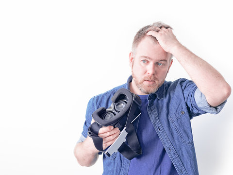 An astonished man in a blue denim shirt holds a game helmet in one hand, holding his head with the other hand. Image on the theme of virtual or augmented reality and computer games