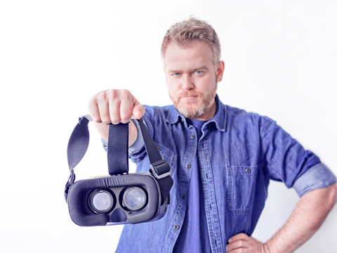 A gloomy red programmer in a blue denim shirt is holding a game helmet in his hand. Image on the theme of virtual or augmented reality, computer games and visual technologies