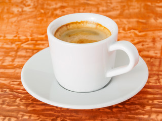 black frothy coffee with foam in white cup with plate on bright copper metal table, side view, close up