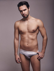one young handsome man, looking at camera, shirtless, fashion model, gray background wearing briefs.