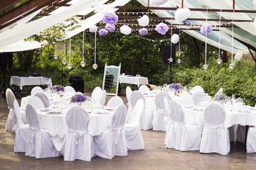 Outdoor wedding in Scandinavian style in old abandoned greenhouse. White tablecloth, white chairs, green trees in background. 