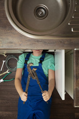woman in blue overalls with adjustable wrench under sink top view