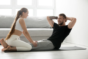 Fit young man doing crunch fitness exercise at home lying on floor with girlfriend supporting helping boyfriend, sporty woman training husband at home, abs abdominal sit ups, love as good motivation