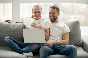 Young happy couple laughing with joy sitting at home on sofa looking at laptop screen, cheerful man and woman watching funny video on computer, having fun enjoying positive amusing content online