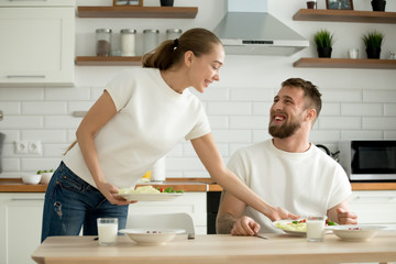 Attractive caring wife serving food cooked for husband in cozy kitchen, grateful man appreciating woman prepared healthy breakfast dinner, happy couple enjoying meal together at home dining table