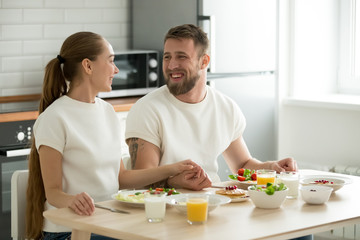 Happy young couple holding hands having breakfast cooked at home sitting at kitchen dining table,...