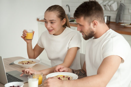 Young Couple Enjoying Meal Talking Using Laptop On Dining Table, Husband And Wife Having Oatmeal Breakfast Looking At Computer While Eating Reading Discussing Online News Or Checking Social Networks