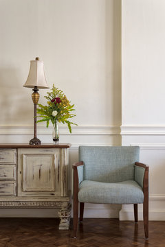 Interior shot of light blue armchair and cream vintage sidebar with table lamp and flowers planter on off white wall and wooden floor