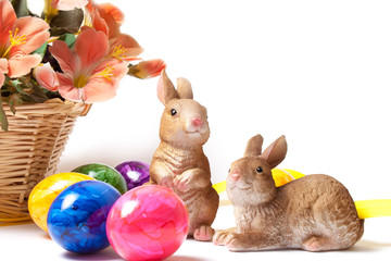 Bunte Ostereier, zwei Osterhasen und gelbes Band mit dekorativen Blumen im Korb auf weißem Hintergrund.