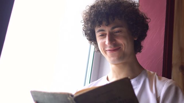 Young Curly Man in White Cloth is Sitting on Window Sill and Reading Book with Daylight from Window