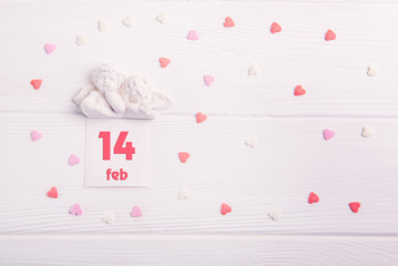 Top view statuette of two little lovely angels with card with 14 febuary date on the white wooden background with multicolor sweet hearts. Valentine's day concept.