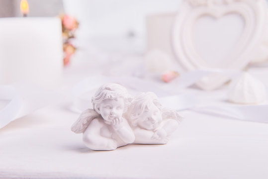 statuette of two antique little lovely angels of the gypsum on the white wooden table with photo frame, candle and flowers. Love background. Copy space, selective focus.