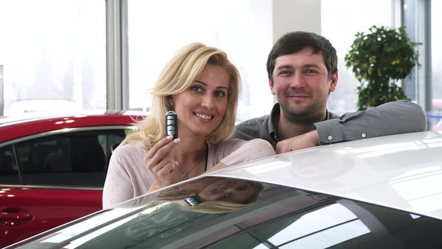 Cheerful mature couple leaning on their new car at the dealership smiling joyfully holding car keys happiness emotions buying consumerism travelling ownership safety retail sales.