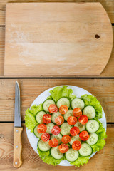 a wooden table is a knife and a cutting board and on a white plate are sliced cucumbers, tomatoes and lettuce leaves