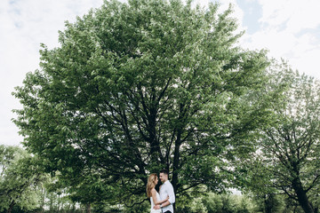 A young guy and an attractive girl hug each other near a large green tree in the garden. Love story