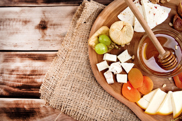 Cheese plate - various types of cheese, honey, grapes, dried apricots, nuts and figs on a wooden board on dark wooden background.