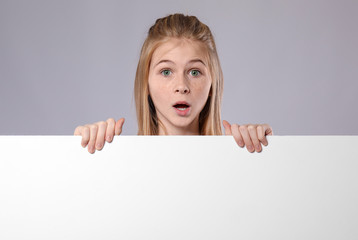 Cute surprised girl with blank advertising board on grey background