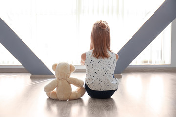 Little girl with teddy bear sitting on floor near window in empty room. Autism concept