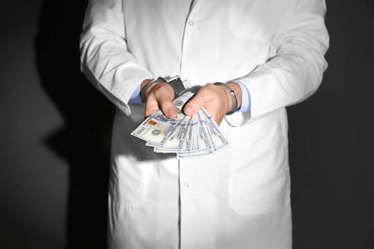Male doctor in handcuffs holding bribe on black background