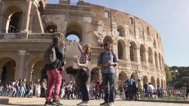 Three young friends tourists standing in front of colosseum in rome reading map guide for directions pointing with backpacks sunglasses happy beautiful girl long hair