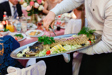 Dish of whole fish on a tray in the hand of the waiter. Male waiter handing a dish, and holding a tray with baked fish and garnish, close-up