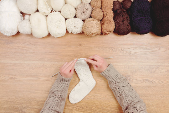 Dressmaker workplace. Woman female girl hands knitting warm white socks by knitted needles on wooden background with line of balls of thread