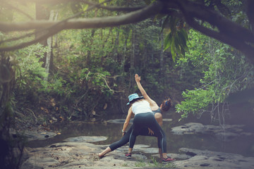 Beautiful mother and daughter are practicing yoga on rocks near the streams, waterfalls. In the rain forest on the mountain. Exercise on vacation Family relationship and nature concept.