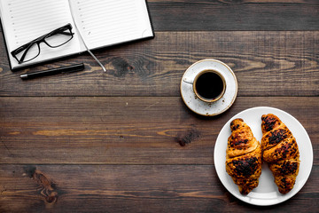 Work at breakfast. Coffee and croissants near notebook and glasses. Dark wooden background top view copy space