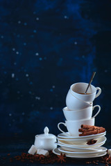 Stack of balancing white coffee cups and tea saucers with coffee beans and cinnamon on a dark blue background. Creative barista concept with copy space.
