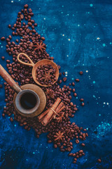 Сopper cezve on scattered coffee beans with handmaiden wooden scoop, cinnamon and anise stars on a dark background. Top view hot drink photography with copy space.