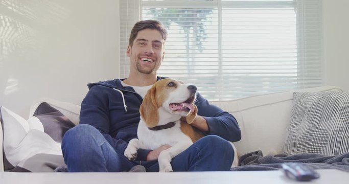 Smiling man sitting on sofa stroking his pet dog while watching television  