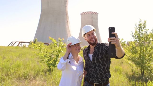 Two engineers in white helmets, a man and woman taking selfies at the construction site on the background of cooling towers.