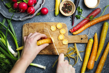 Woman hands cutting carrots on wooden board and fresh spring vegetables for vegetarian cooking on the table, top view