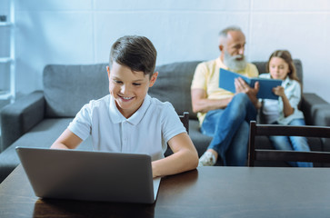 Favorite leisure activity. Waist up shot of a joyful little boy smiling while spending his leisure time at a laptop with his grandfather and sister reading a book in the background.