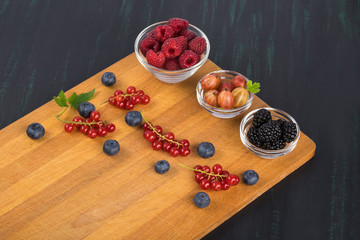 berries raspberries, gooseberries and blackberries in glass bowls stand on a cutting board with berries of red currants and blueberries, which lies on a black table