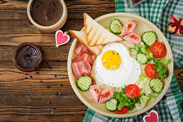 Breakfast on Valentine's Day - fried egg in the shape of a heart, toasts, sausage, bacon and fresh vegetables. English breakfast. Cup of coffee. Top view