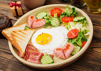 Breakfast on Valentine's Day - fried egg in the shape of a heart, toasts, sausage, bacon and fresh vegetables. English breakfast