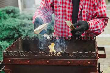 man in plaid shirt puts planks in fire of barbecue