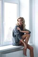 Young beautiful blonde is sitting on window in gray sweater