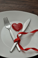 A fork an knife tied together with a red ribbon displayed on a white plate with a red heart shape lollipop