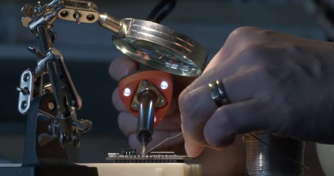 Hobbyists' hands soldering an electrical connection on circuit board with a hot soldering iron fitted with LED lights. Magnifying glass and clamp visible. Recorded in 4K, slow motion at 60fps.