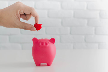 Man's hand putting red heart in to piggy bank metaphor saving love for lover or family in every...